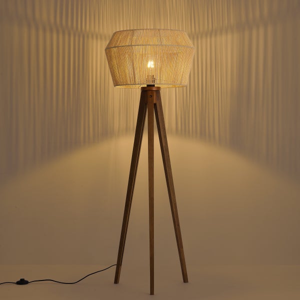 Dreibein-Stehleuchte aus Holz mit rundem Seilschirm, eingeschaltet mit warmem Licht und Streifenschatten an der Wand.