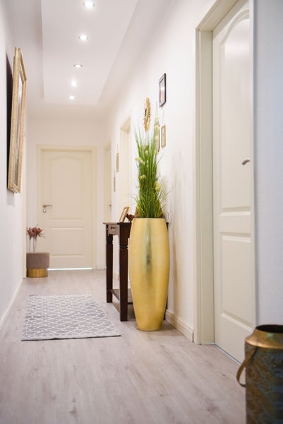 Heller, organisierter Flur mit weißen Innentüren, Laminatboden in Holzoptik, einem grauen Läufer und einer großen goldenen Bodenvase mit Kunstpflanzen.