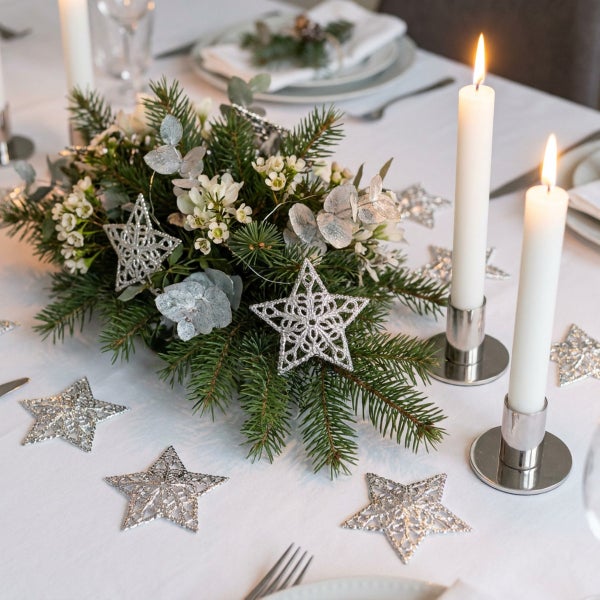 Weihnachtliche Tischdekoration mit Tannengrün, weissen Blumen, glitzernden Sternen und zwei brennenden Stabkerzen in modernen Kerzenhaltern.