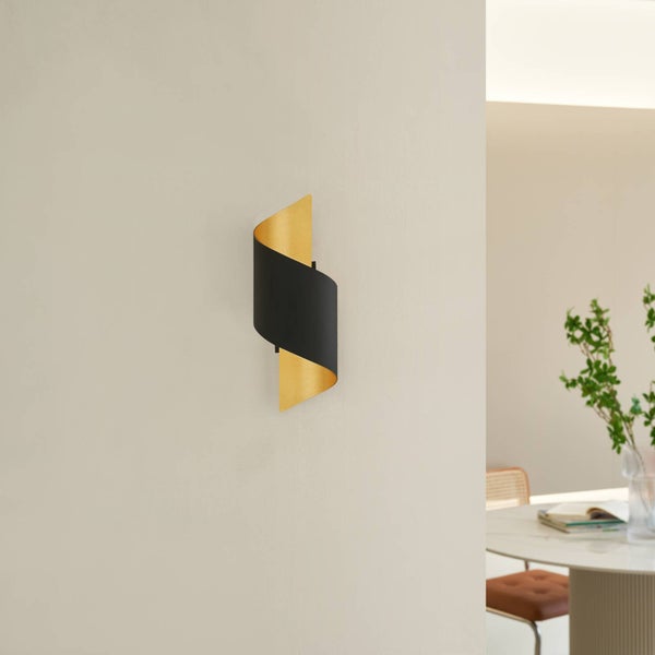 Moderne Wandleuchte mit geschwungenem Metallschirm in Schwarz und Gold an einer hellen Wand in einem hellen und organisierten Raum.