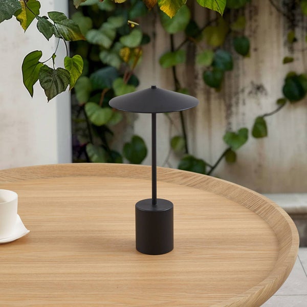 Moderne schwarze LED-Außentischleuchte mit schirmförmigem Schirm auf einem Holztisch vor einer Wand mit grünen Kletterpflanzen.