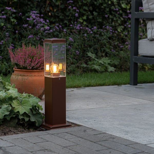 Moderne Außenleuchte in Säulenform auf einer Terrasse mit warmem Licht, umgeben von Pflanzen.