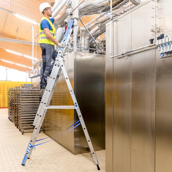 Ein Mann mit Schutzhelm und Warnweste steht auf einer Zarges Skymaster Leiter in einer industriellen Umgebung.