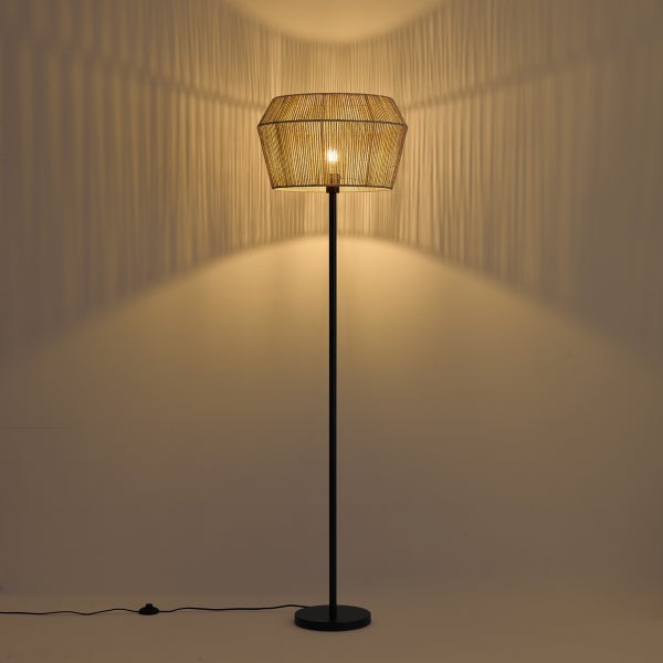 Moderne Stehlampe mit schwarzem Metallgestell und naturfarbenem, gewebtem Schirm, die ein warmes Licht und Streifenmuster an die Wand wirft.