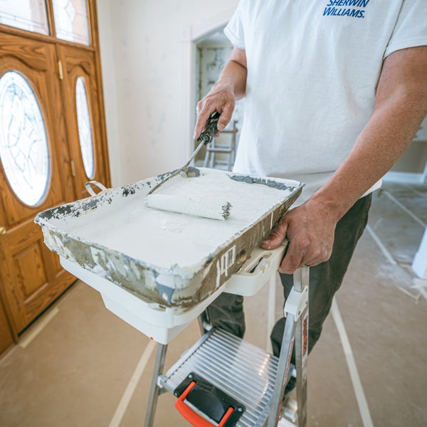 Maler mit Sherwin-Williams Logo auf einer Trittleiter taucht eine Farbwalze in eine Farbwanne mit weißer Farbe.