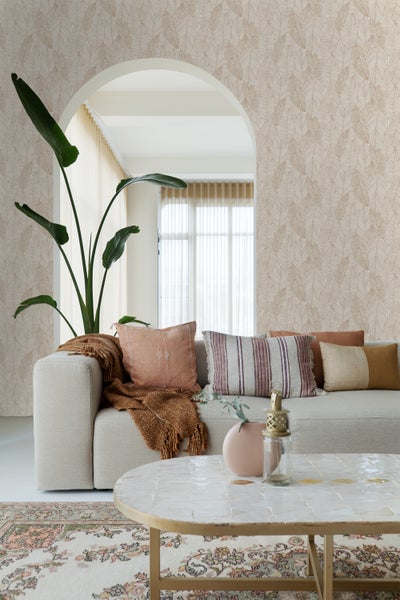 Wohnzimmer mit beiger Couch, Kissen, Decke, Tisch und Blattmuster-Tapete für eine helle und organisierte Atmosphäre.