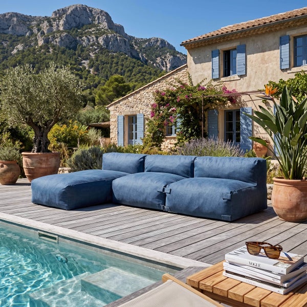 Modulares blaues Gartensofa auf einer Holzterrasse am Pool, im Hintergrund ein mediterranes Steinhaus und Berge, helle Atmosphäre.