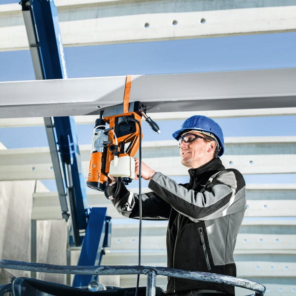 Ein Arbeiter mit blauem Helm und Schutzbrille bedient eine Magnet-Kernbohrmaschine an einem weißen Stahlträger auf einer Baustelle.