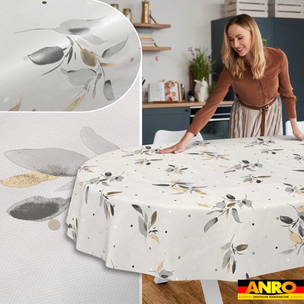 Frau breitet eine runde Tischdecke mit Blättermotiv auf einem Esstisch aus. ANRO Deutsche Manufaktur Logo.