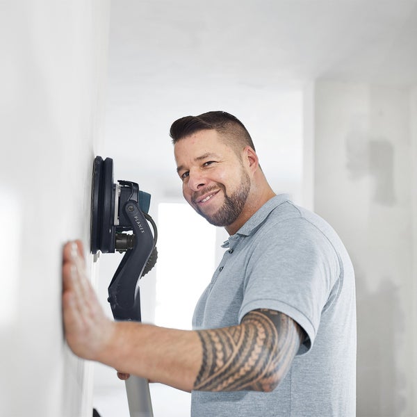 Handwerker nutzt einen Langhalsschleifer Planex zum Schleifen einer weißen Wand in einem hellen, renovierten Innenraum.