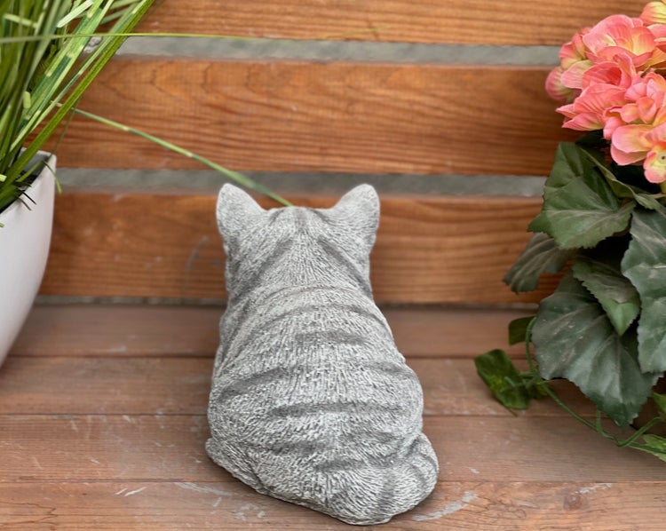 Gartenfigur in Form einer sitzenden Katze von hinten, in Steinoptik, dekoriert auf einer Holzbank neben Blumen.