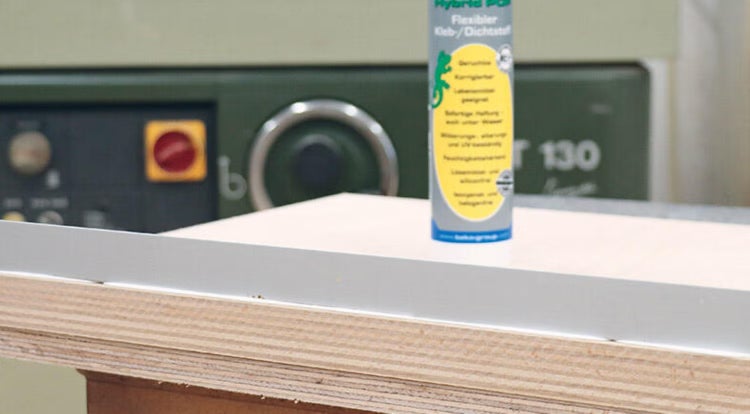 Kartusche mit flexiblem Kleb- und Dichtstoff auf einer Arbeitsplatte neben einer Holzplatte mit Profilleiste in einer Werkstatt.