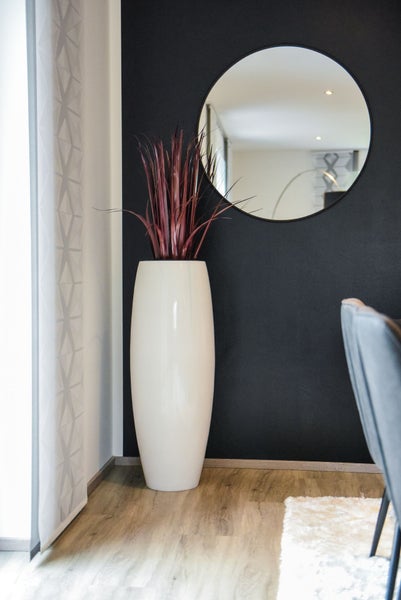 Hohe weiße Bodenvase mit roten Ziergräsern vor einer dunklen Wand mit rundem Spiegel und hellem Holzboden.