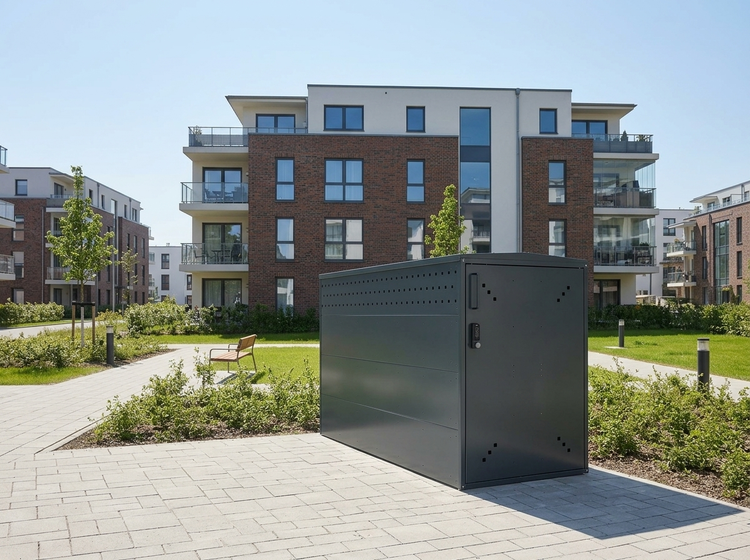 Dunkelgraue Fahrradgarage aus Metall vor einem modernen Wohnhaus auf einer gepflasterten Fläche.