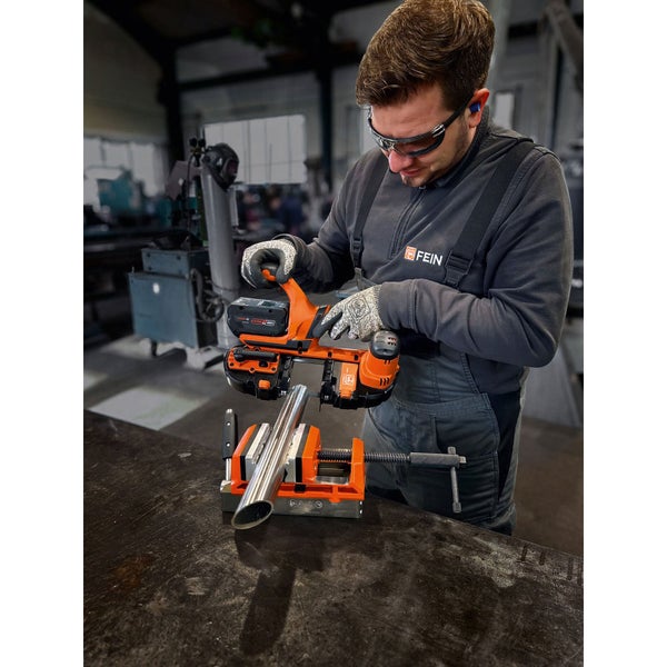 Ein Mann benutzt eine Akku-Bandsäge von Fein, um ein Metallrohr zu schneiden, das in einem Schraubstock fixiert ist. Er trägt Schutzkleidung und Gehörschutz.