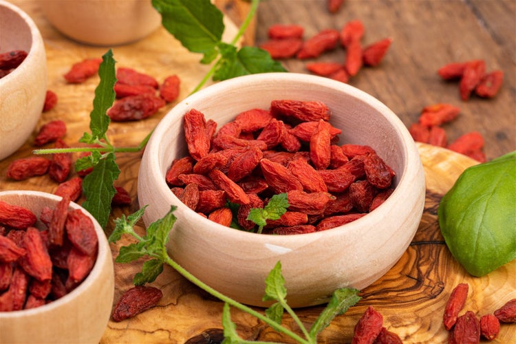 Getrocknete Goji-Beeren in Holzschalen auf einer Holzoberfläche mit grünen Blättern.