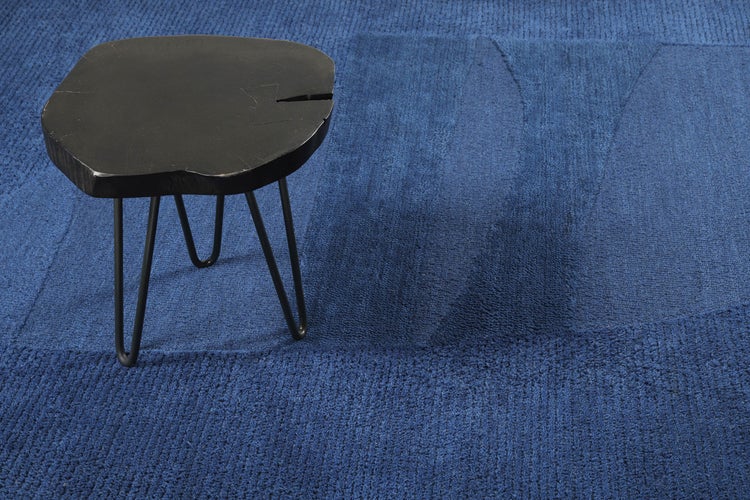 Blauer Teppich mit geometrischem Reliefmuster und abgerundeter Form in einem hellen Wohnzimmer vor einem weißen Sofa.