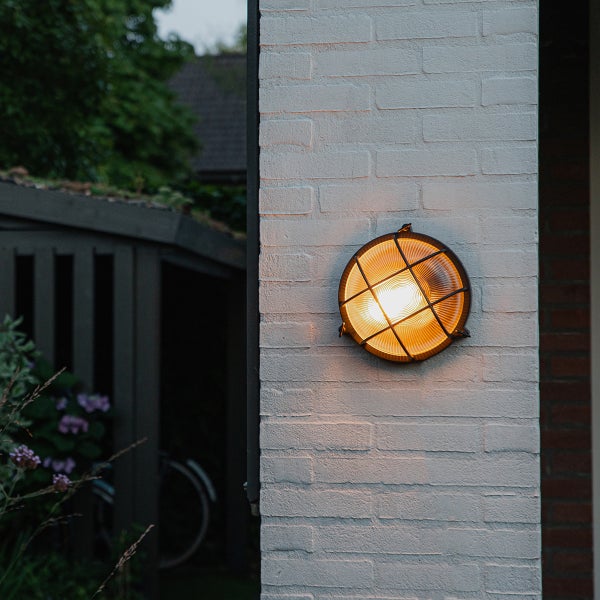 Runde Außenwandleuchte mit Metallgitter, leuchtet mit warmem Licht an einer weißen Backsteinwand.