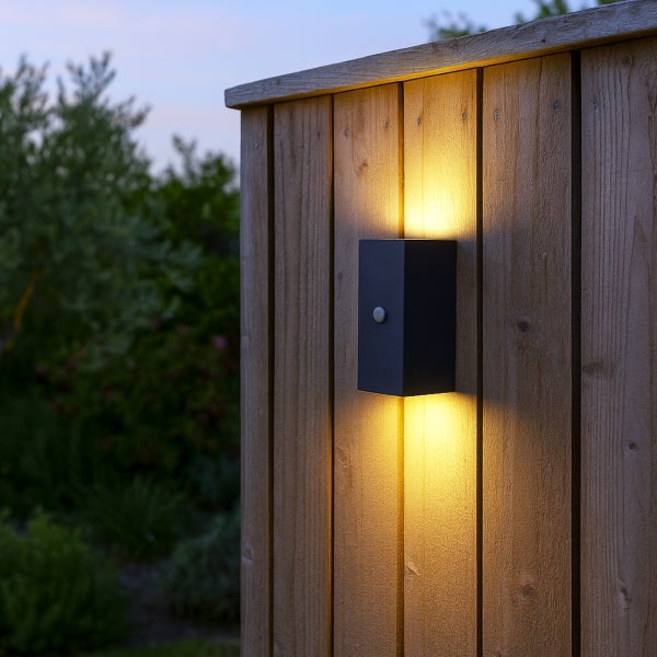 Moderne anthrazitfarbene LED-Außenwandleuchte mit Licht nach oben und unten sowie Bewegungsmelder montiert an einer Holzwand im Garten.
