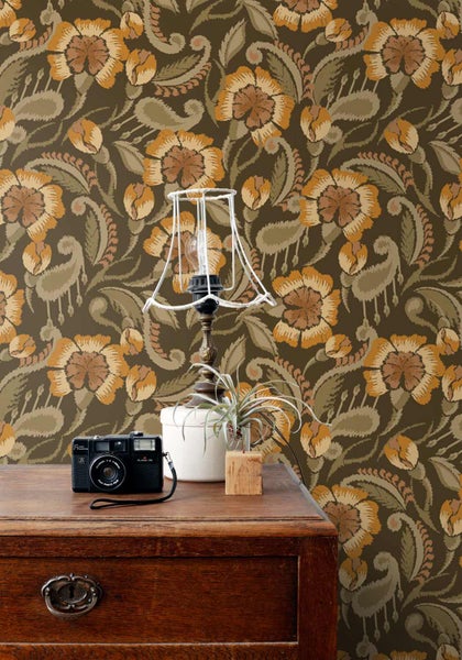 Tapete mit floralem Retromuster hinter einer Holzkommode mit Kamera und Drahtlampe.