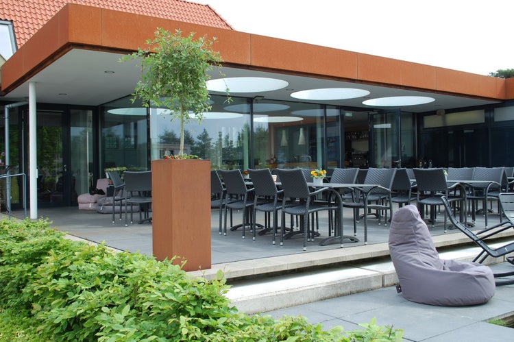 Moderne Außenterrasse mit grauen Gartenmöbeln, einem hohen Pflanzgefäß aus Cortenstahl und einem Gebäude mit großen Glasfronten.