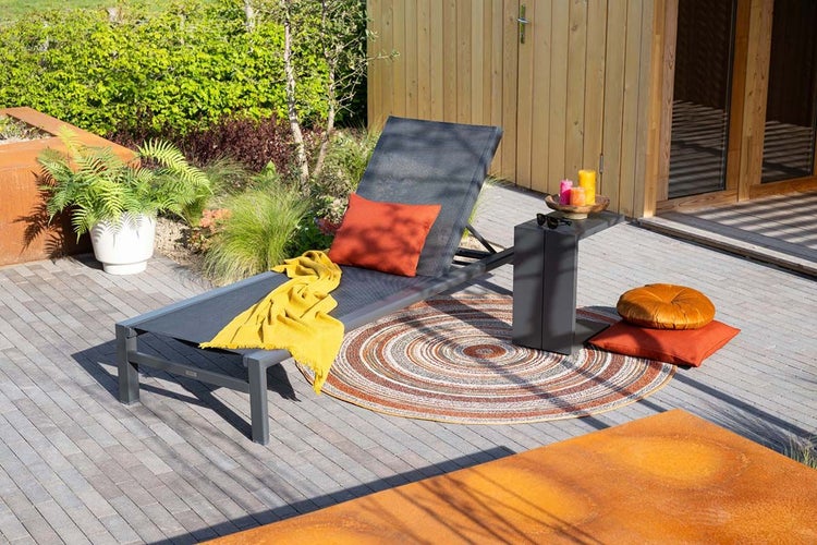 Moderne Gartenliege in Anthrazit auf gepflasterter Terrasse mit gelber Decke, orangefarbenem Kissen, Beistelltisch und rundem Teppich.