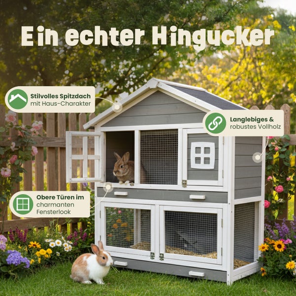 Zweistöckiger Kaninchenstall aus massivem Vollholz in Grau und Weiß mit Spitzdach, Gitterfenstern und Kaninchen in einem Garten.