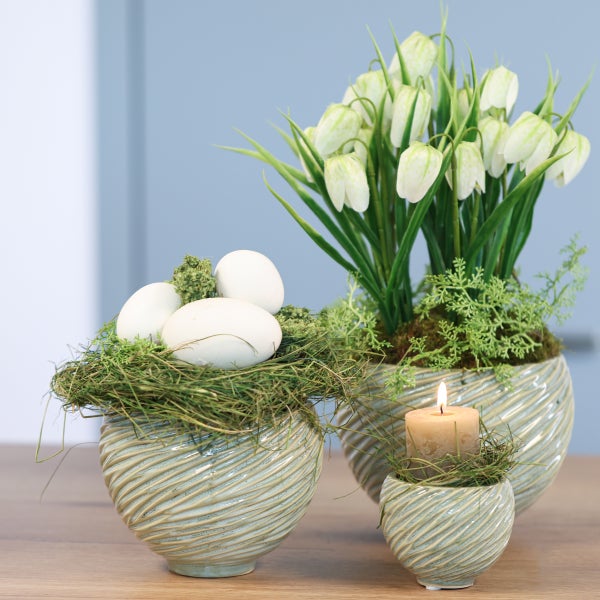 Frühlingshafte Osterdekoration in strukturierten Keramiktöpfen mit Ostereiern, einer brennenden Kerze und Glockenblumen auf einem Holztisch.