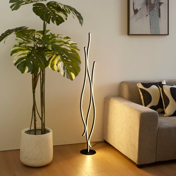 Moderne wellenförmige LED-Stehlampe im Wohnzimmer neben Monstera-Pflanze und Sofa, helles und geordnetes Ambiente mit warmem Licht.