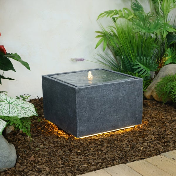 Moderner, quadratischer Gartenbrunnen in Steinoptik mit Wasserspiel und LED-Beleuchtung am Sockel, platziert auf Rindenmulch vor Pflanzen.