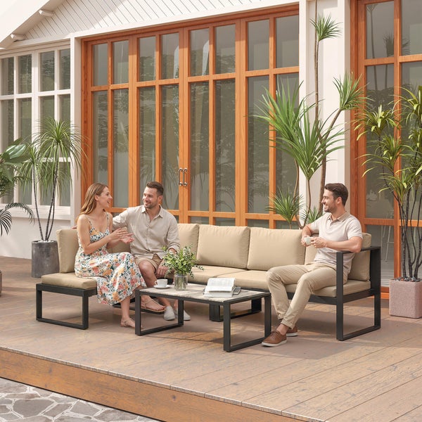 Modernes Garten-Lounge-Set auf einer Holzterrasse mit einem modularen Sofa, beigen Polstern und passendem Couchtisch vor einer Fensterfront.