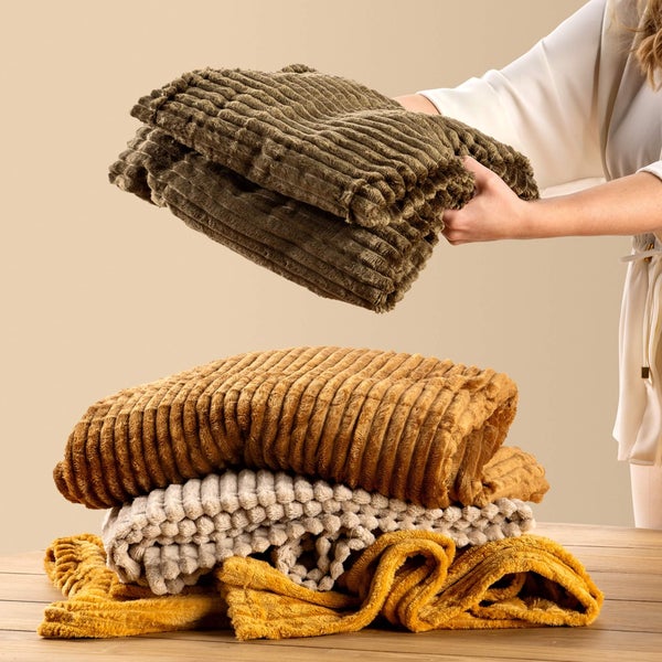 Person hält eine gefaltete strukturierte Wohndecke über einen Stapel ähnlicher gerippter Decken in verschiedenen Farben auf einem Holztisch.