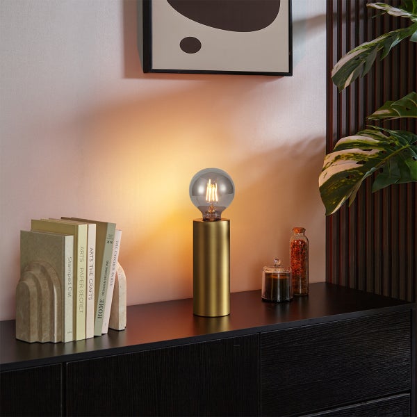 Moderne goldene Tischleuchte mit runder Glühbirne auf einer schwarzen Kommode neben Büchern und Dekoration.