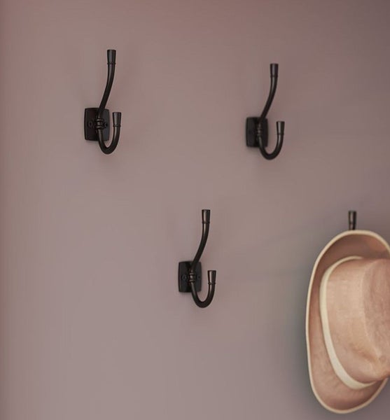 Mehrere an der Wand montierte Garderobenhaken aus Metall mit zwei Hakenarmen, einer davon trägt einen Hut.