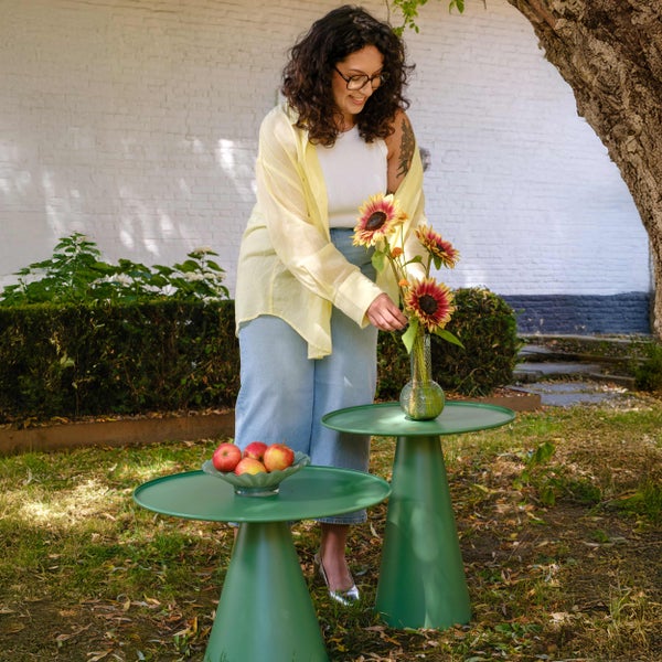 Frau arrangiert Sonnenblumen auf einem grünen runden Gartenbeistelltisch aus Metall neben einem zweiten Tisch mit Äpfeln in einem hellen Garten.