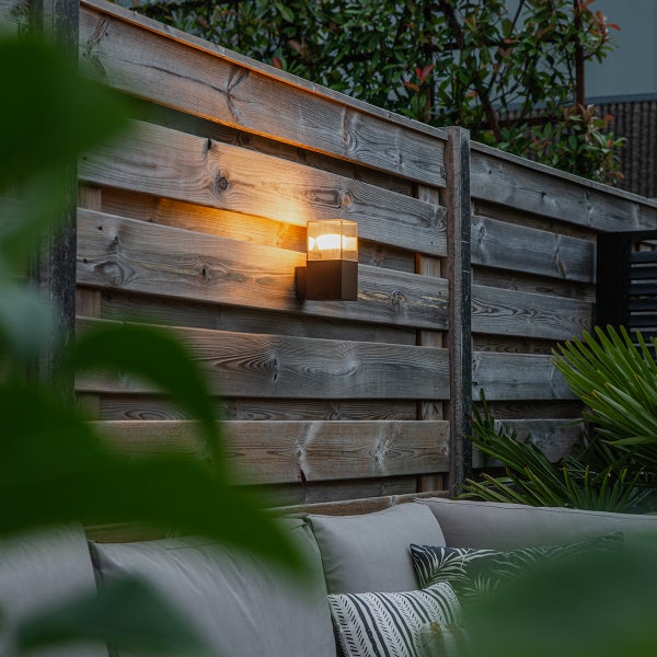 Moderne Außenwandleuchte an einem Holzzaun montiert, beleuchtet einen Sitzbereich im Garten mit Sofa und Kissen.