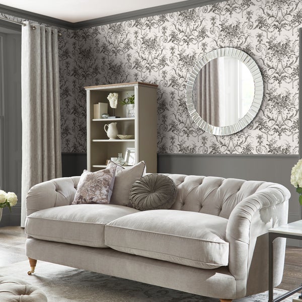 Wohnzimmer mit barocker Tapete mit Blumenmuster, beige gepolstertem Sofa, rundem Wandspiegel und offenem Regal mit Dekorationsgegenständen.