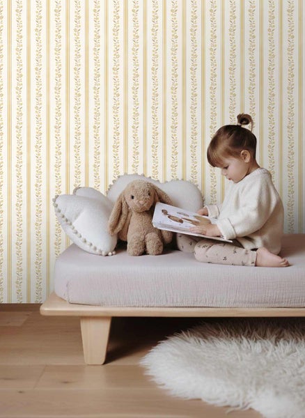 Vliestapete mit gelben vertikalen Streifen und feinen Blumenranken in einem hellen Kinderzimmer mit Holzbett und Stofftieren.