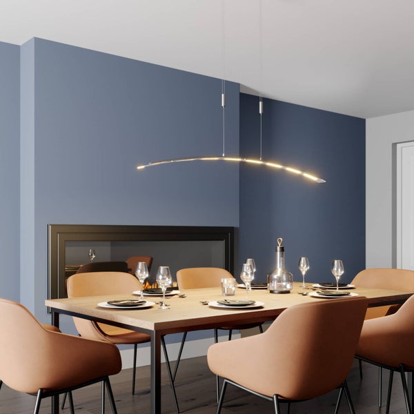 Moderne, gebogene LED-Pendelleuchte über einem gedeckten Holztisch in einem Esszimmer mit Kamin und blauen Wänden.