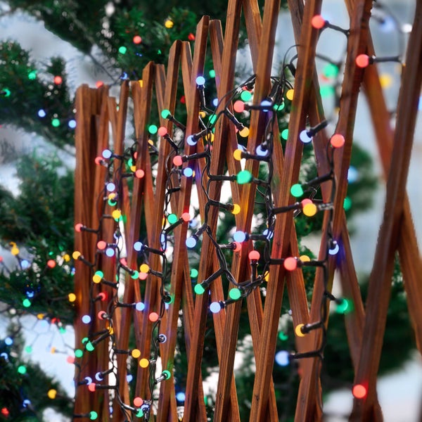 Bunte LED-Lichterkette dekoriert an einem hölzernen Rankgitter vor grünen Pflanzen.