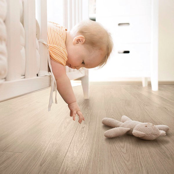 Ein Kleinkind greift aus einem Kinderbett nach einem Stofftier auf einem hellen Holzboden in einem modern eingerichteten Zimmer.