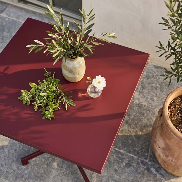 Quadratischer Gartentisch aus Metall in Bordeaux auf einer Terrasse mit Olivenzweigen in einer Vase und Kräutern dekoriert.