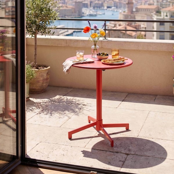 Runder Bistro-Tisch aus Metall in Rot auf einem hellen Balkon mit Blick auf den Hafen, gedeckt mit Getränken und Snacks.