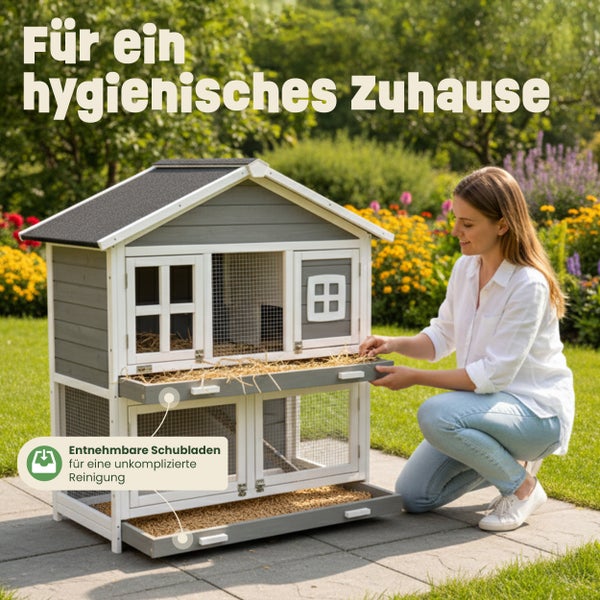 Zweistöckiger Kleintierstall aus grauem und weißem Holz im Garten mit einer Frau, die eine entnehmbare Schublade zur Reinigung herauszieht.
