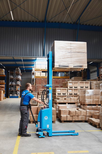 Mitarbeiter bedient einen Elektro-Hochhubwagen in einem Lager und hebt eine mit Kartons beladene Europalette an.
