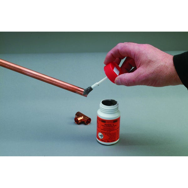 Eine Hand trägt mit einem Pinsel Weichlötpaste auf das Ende eines Kupferrohrs auf, neben einer Dose Lötpaste und einem Kupfer-T-Stück.