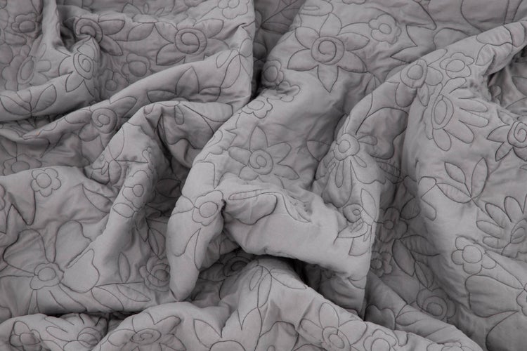 Graue gesteppte Tagesdecke mit Blumenmuster auf einem Bett mit weißen Kissen, daneben eine dekorative Vase und Schale.