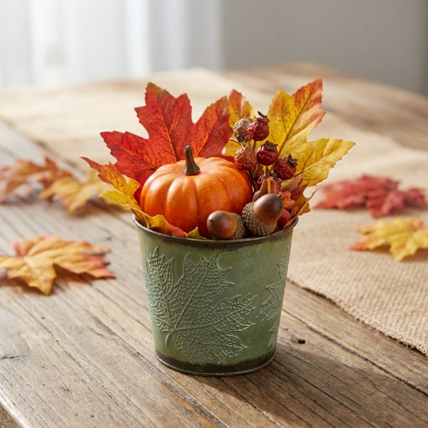 Herbstliche Tischdekoration mit künstlichem Kürbis, Eicheln und Ahornblättern in einem metallenen Übertopf mit Blattprägung.