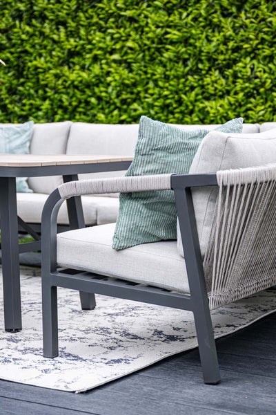 Modernes Garten-Lounge-Set aus Aluminium mit Seilbespannung, hellgrauen Polstern und einem Outdoor-Teppich vor einer Hecke.