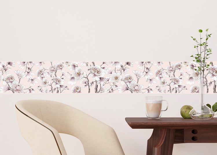 Selbstklebende Tapetenbordüre mit floralem Magnolienmuster an einer hellen Wand in einem Raum mit Holztisch und gepolstertem Sessel.